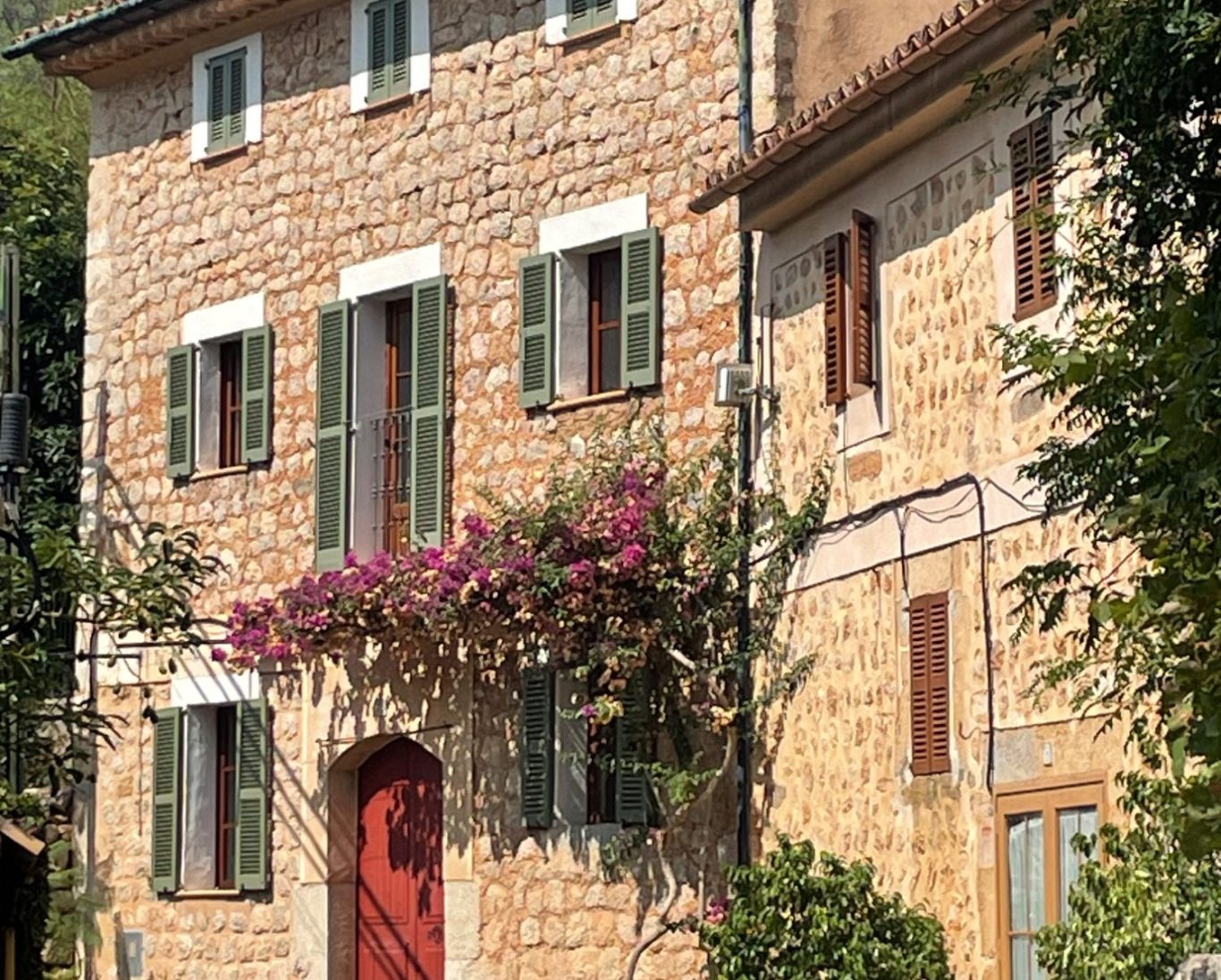 Stadthaus Mallorca - Homeservice - Hausbetreuung Steinernes Haus mit grünen Fensterläden und blühender Pflanze über der Tür.